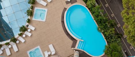 An outdoor swimming pool with lounge chairs and a hot tub, surrounded by greenery and a tiled deck.