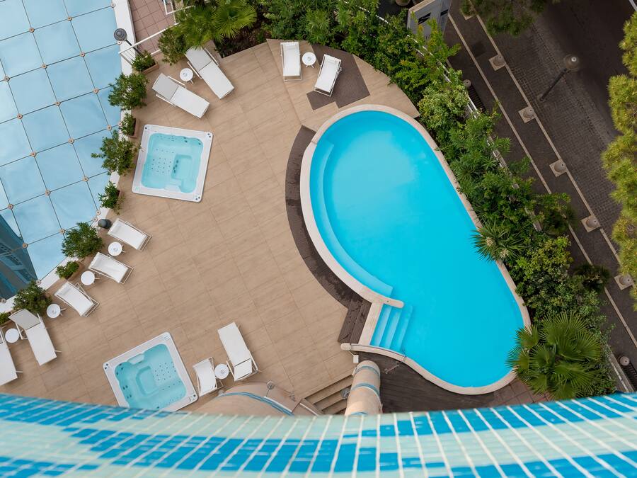 An outdoor swimming pool with lounge chairs and a hot tub, surrounded by greenery and a tiled deck.