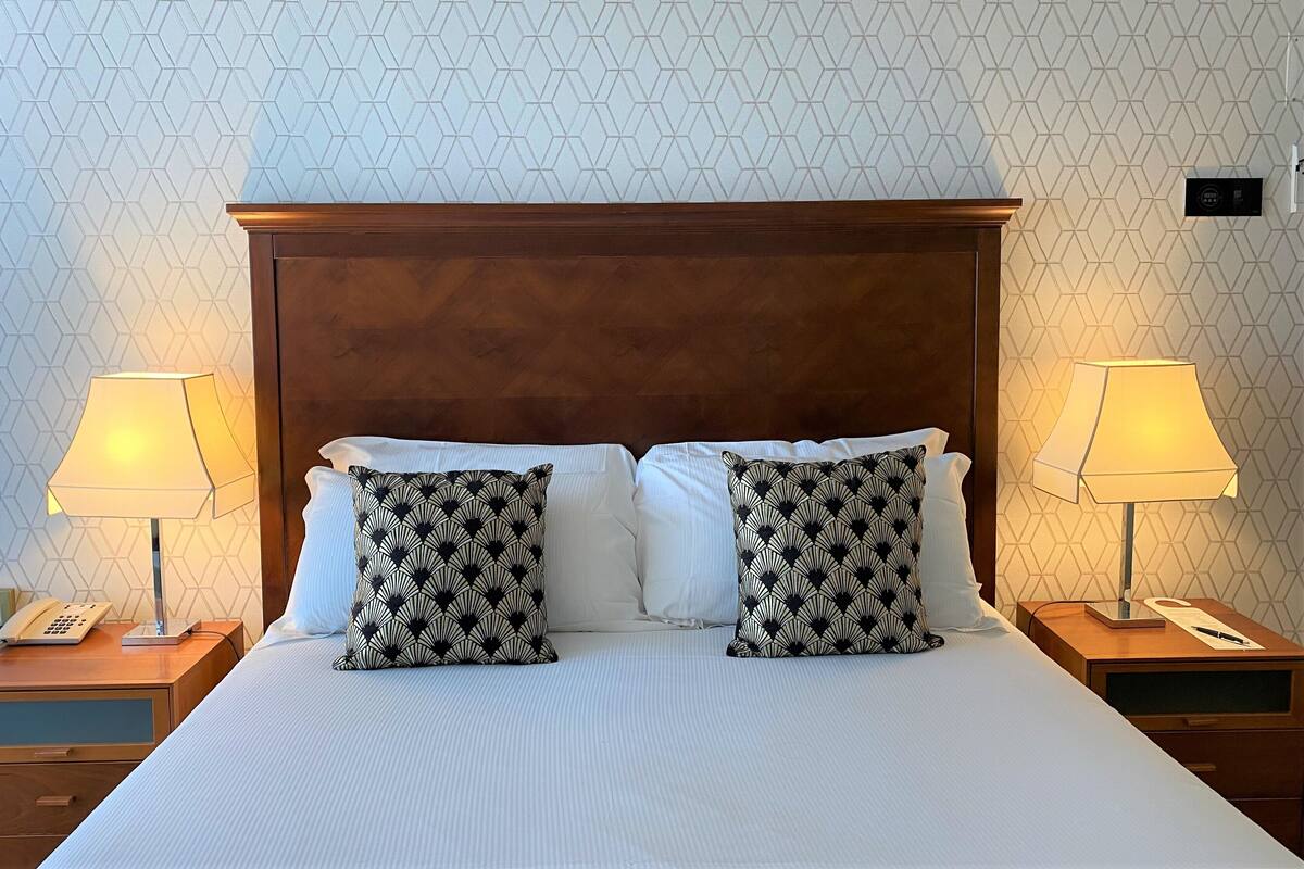 A hotel room with a large bed, two bedside lamps, a wooden headboard, and patterned pillows.