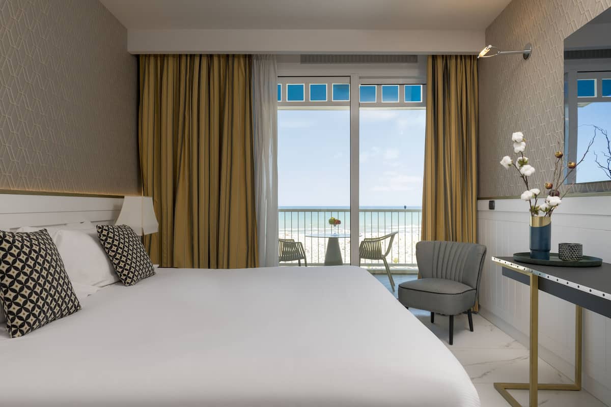 A hotel room with a large bed, a desk with a vase of flowers, a chair, and a view of the beach through a glass door.