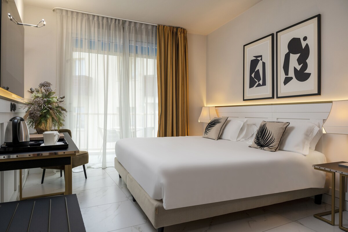Een moderne hotelkamer met een groot bed, een bureau met waterkoker, een kamerplant en abstracte kunst aan de muur.