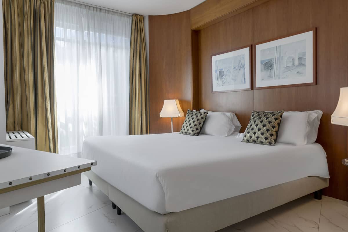 A hotel room with a large bed, a desk, a lamp, and two framed pictures on the wall.