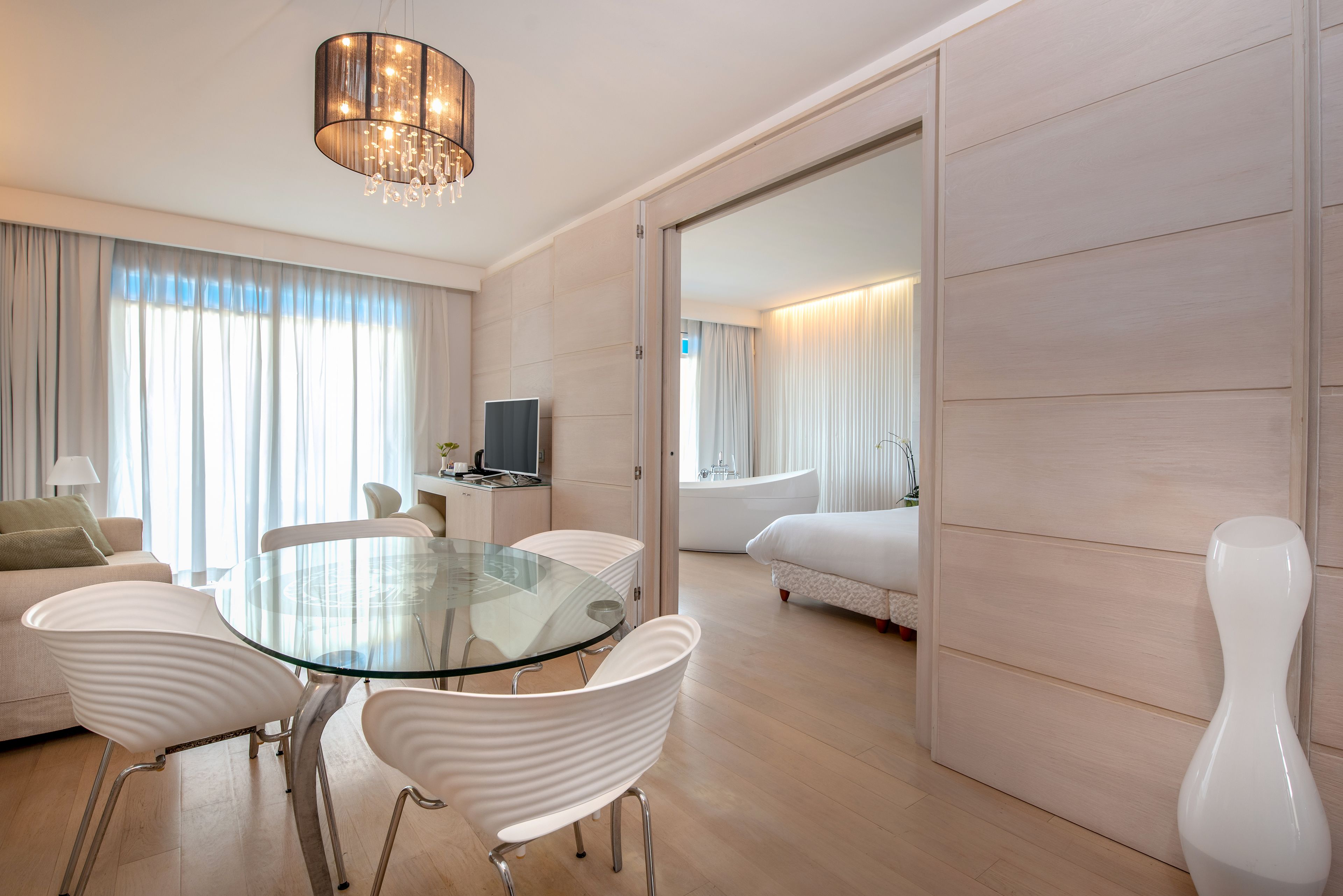 Une chambre d’hôtel moderne avec une table à manger en verre, des chaises blanches, un canapé, un téléviseur et un grand miroir.