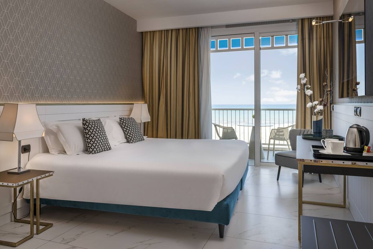 A hotel room with a large bed, a desk with a kettle, and a view of the ocean.