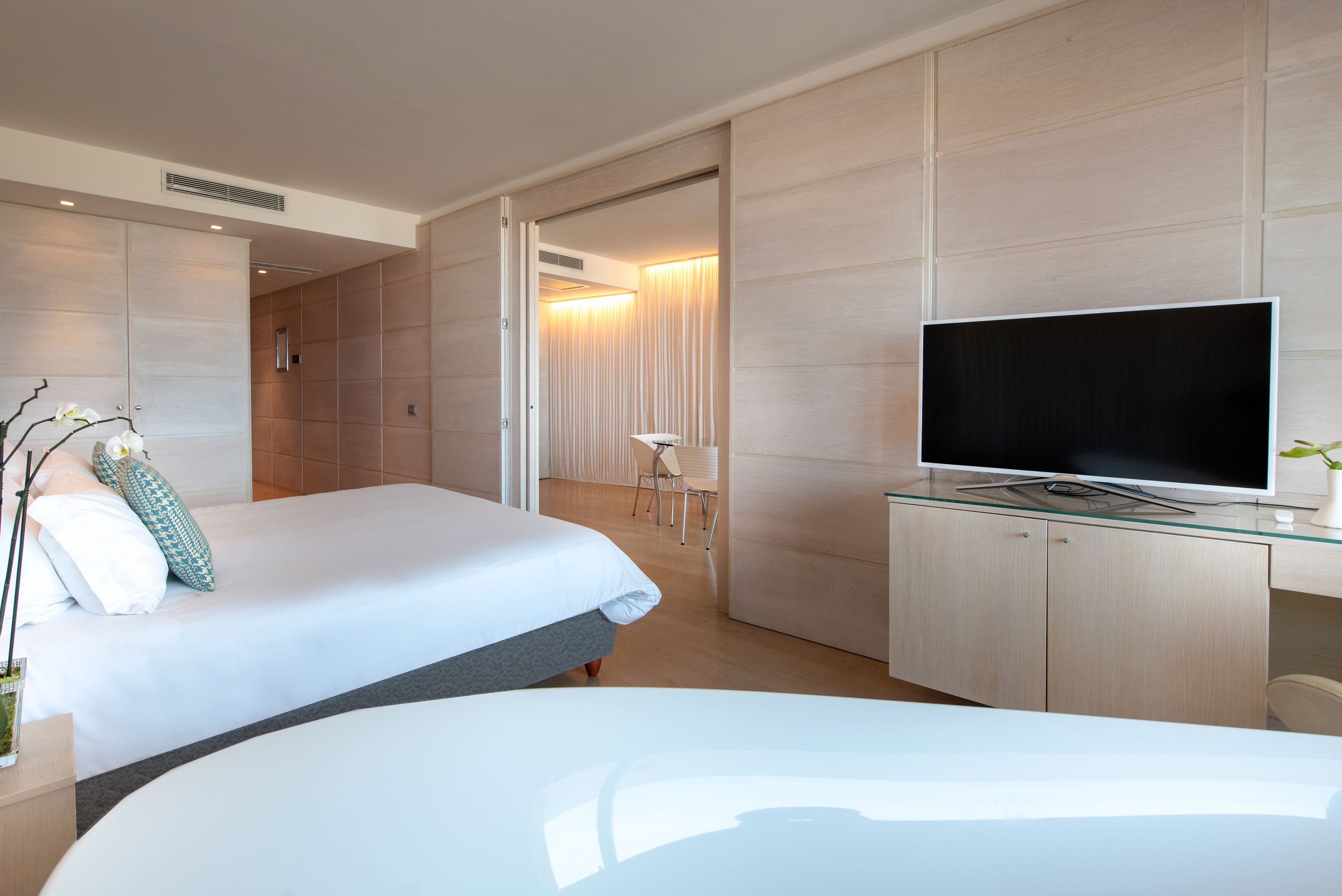 Quarto de hotel moderno com uma cama grande, uma televisão sobre um móvel de vidro e uma área de estar separada com uma cadeira e uma mesa.