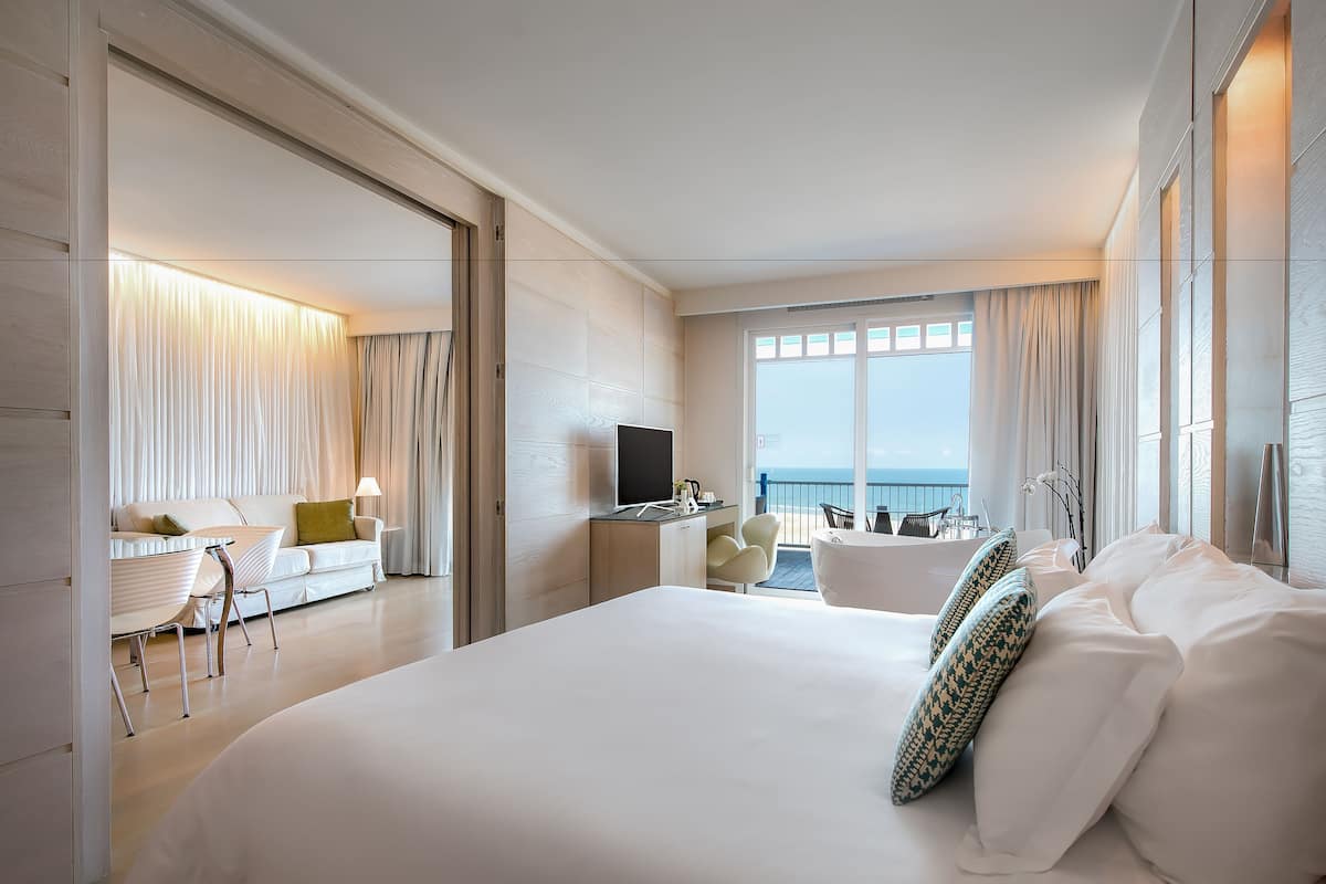 A modern hotel room with a large bed, a desk with a flat-screen TV, a seating area with a sofa and chairs, and a balcony with a view of the sea.
