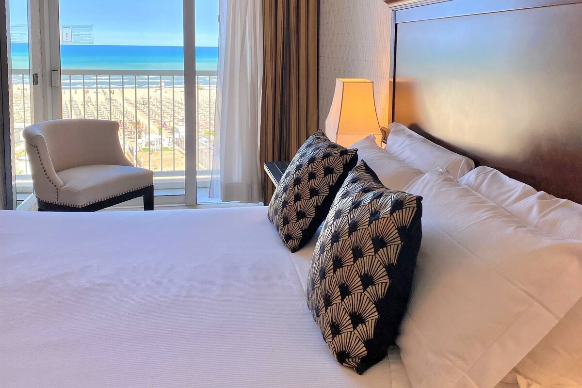 A hotel room with a large bed, two patterned pillows, a chair, and a view of the beach.