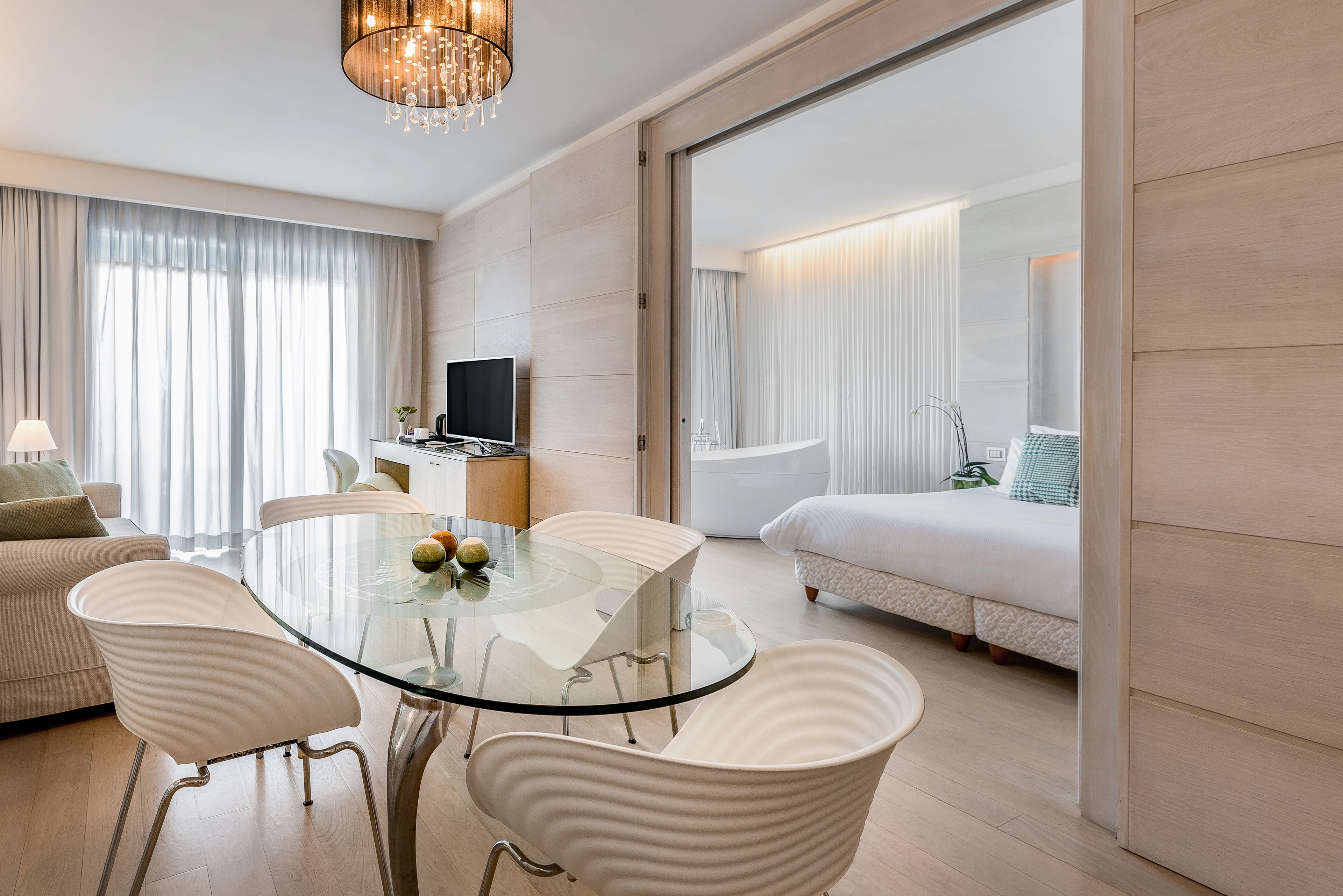 Habitación de hotel moderna con una mesa de comedor de vidrio, sillas blancas tapizadas, un televisor de pantalla plana, un sofá y una cama con edredón blanco.