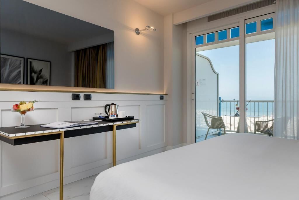 A hotel room with a bed, a desk with a coffee pot, a laptop, and a view of the sea through large windows.