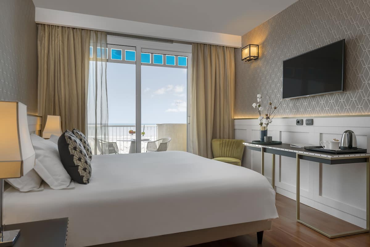 A hotel room with a large bed, a desk with a flat-screen TV, a chair, and a view of the sky through the window.
