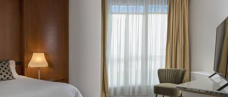 Une chambre d’hôtel avec un lit, un bureau, une chaise, une lampe et une grande fenêtre avec des rideaux.