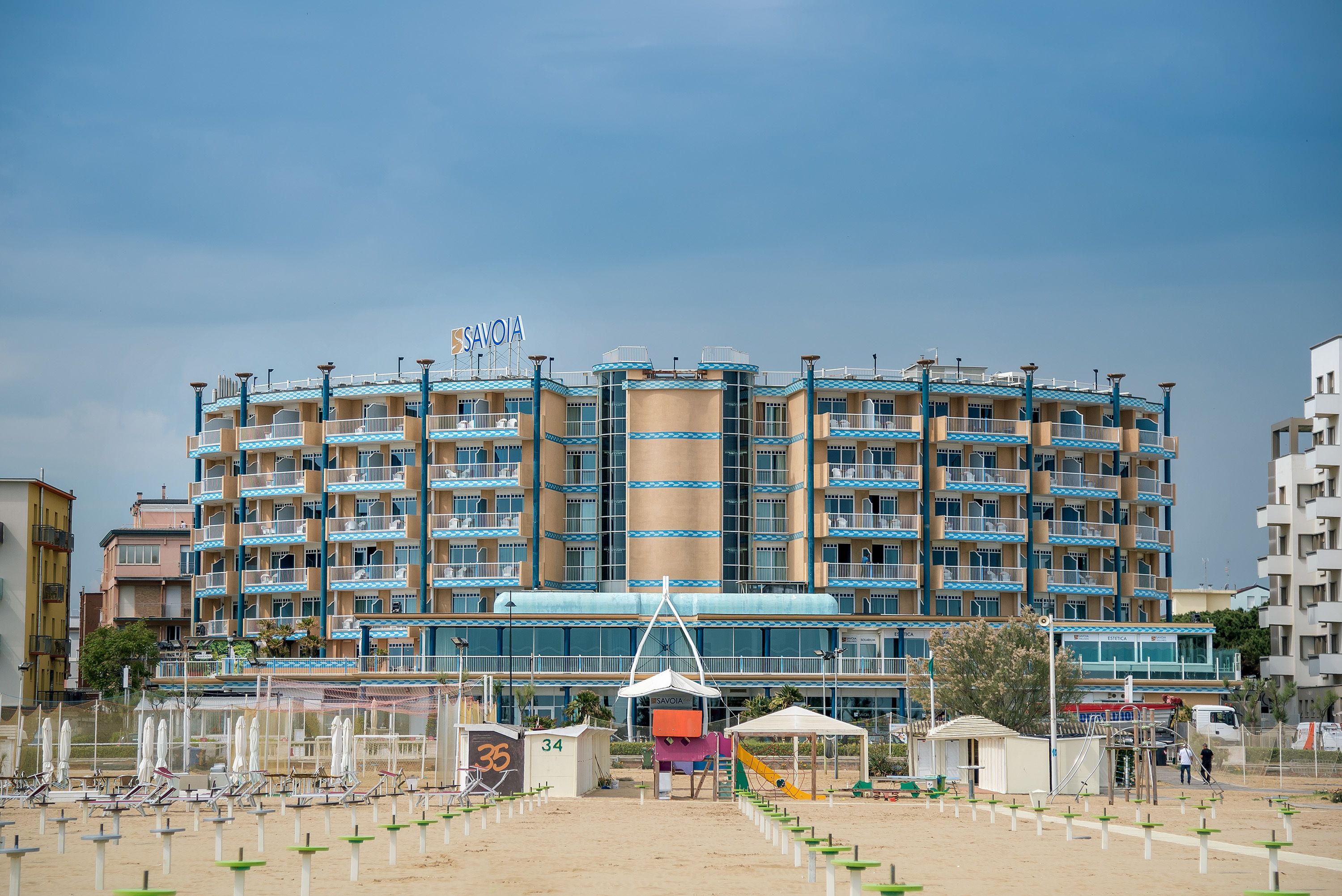 Un grand hôtel doté de nombreux balcons, d’une tour centrale et d’un espace de plage aménagé avec des parasols et des chaises longues.