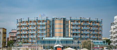 Un grand hôtel doté de nombreux balcons, d’une tour centrale et d’un espace de plage aménagé avec des parasols et des chaises longues.