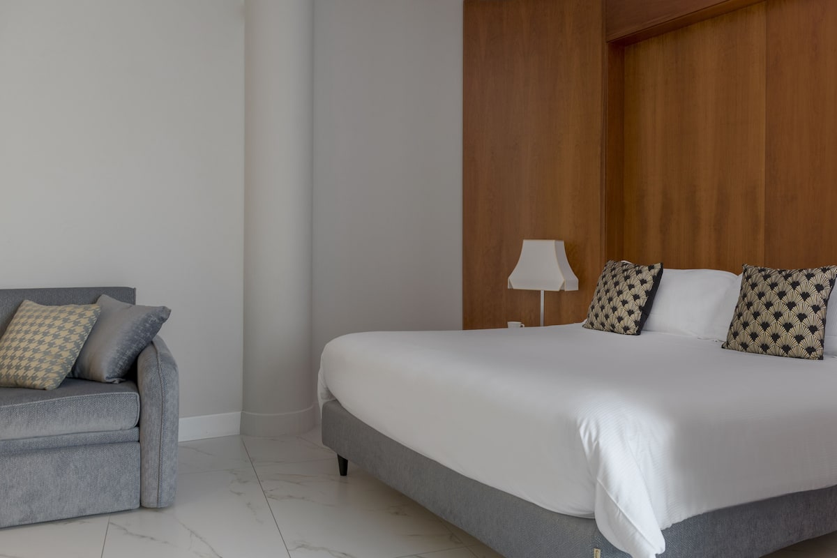 A bedroom with a bed, a grey sofa, and a wooden headboard.