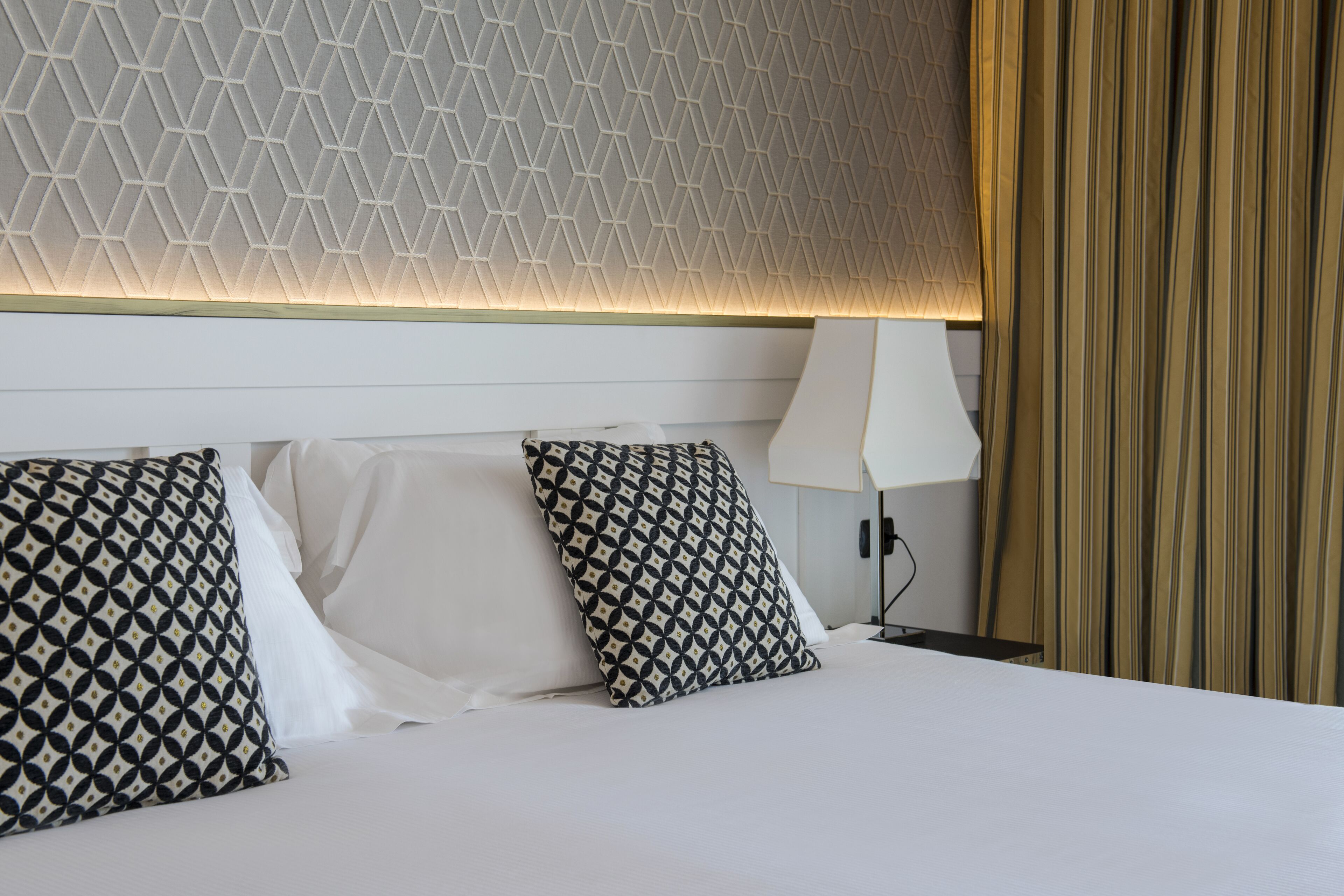 Habitación de hotel con una cama bien tendida, almohadas con estampado geométrico, una lámpara de noche y un techo con textura.
