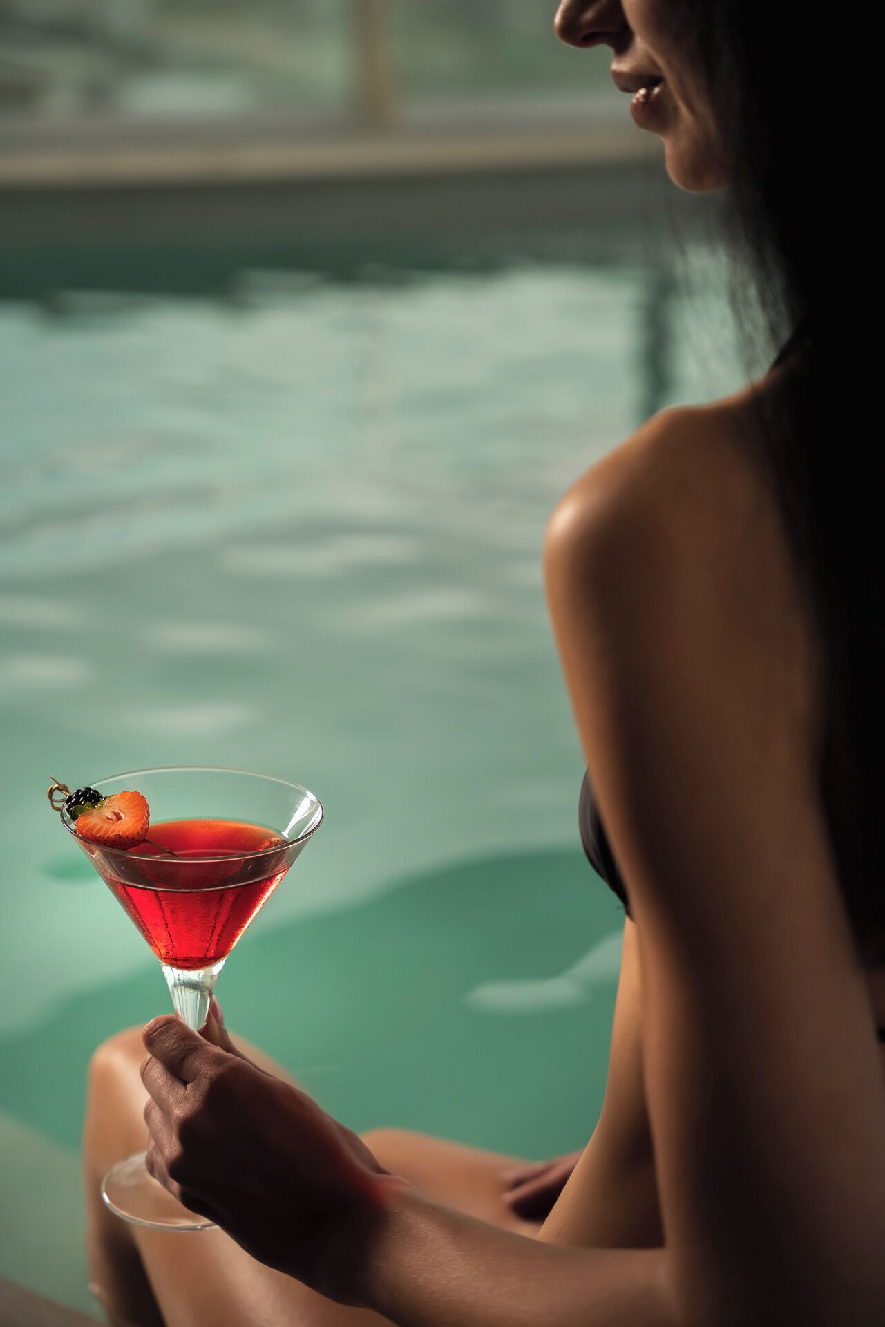 A person holding a martini glass with a cherry and a blackberry garnish, sitting by a pool.