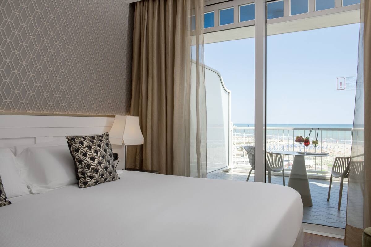 A hotel room with a large bed, a view of the beach, and a balcony with a table and chairs.