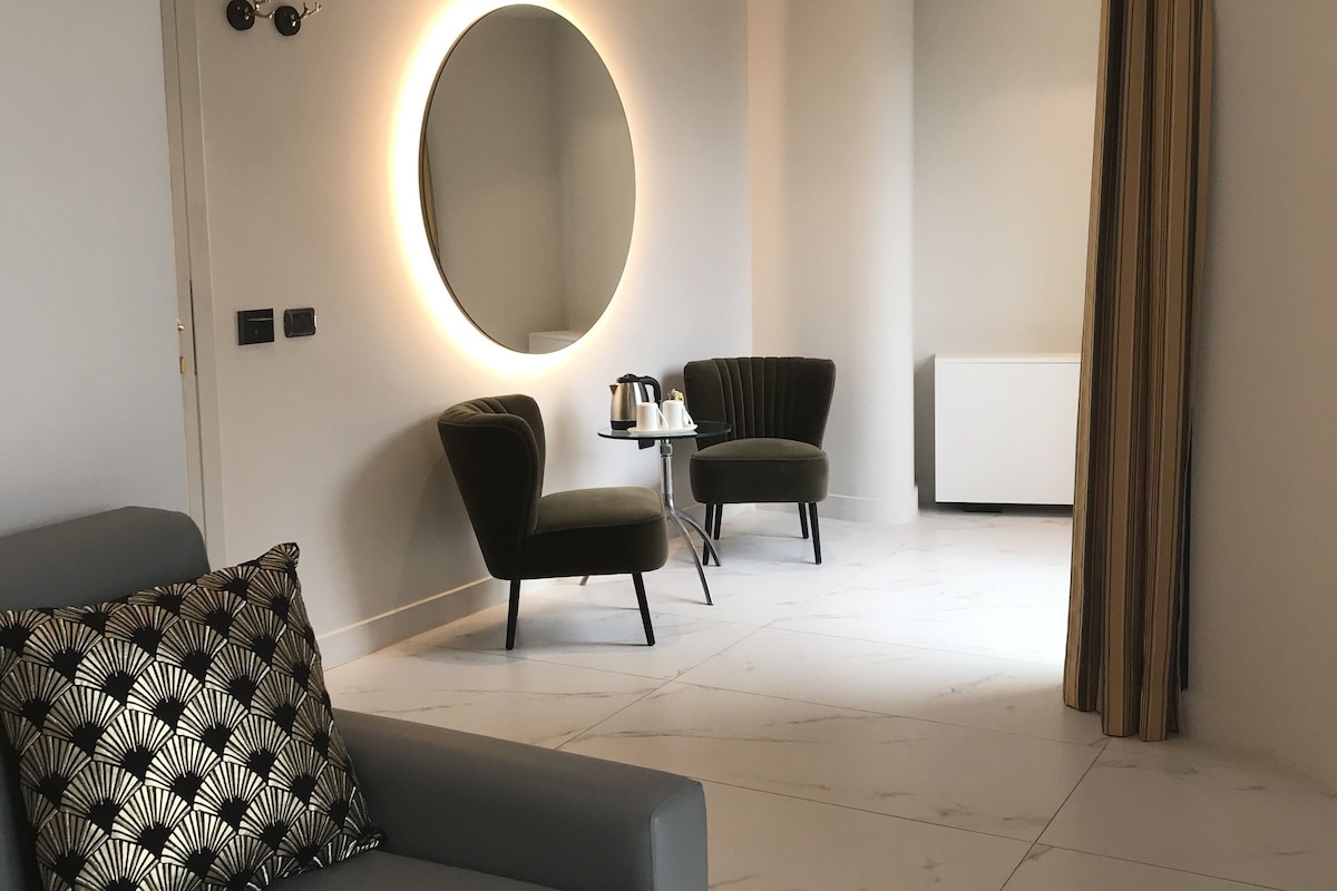 Een moderne, minimalistisch ingerichte kamer met een grijze bank, een ronde spiegel met ingebouwde verlichting, twee donkere fauteuils, en een klein tafeltje met waterkoker en kopjes.