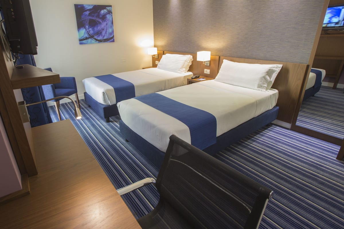 A hotel room with two beds, a desk, and a chair. There is a TV on the desk and a painting on the wall.