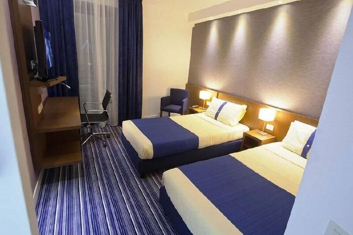 A hotel room with two beds, a desk, a chair, a TV, and a large window with curtains.