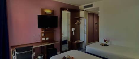 A hotel room with two beds, a desk with a flat-screen TV, a mirror, and a small kitchenette area.