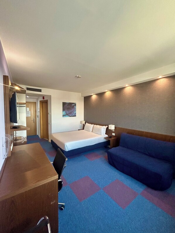A hotel room with a bed, a desk, a chair, a sofa, and a television.