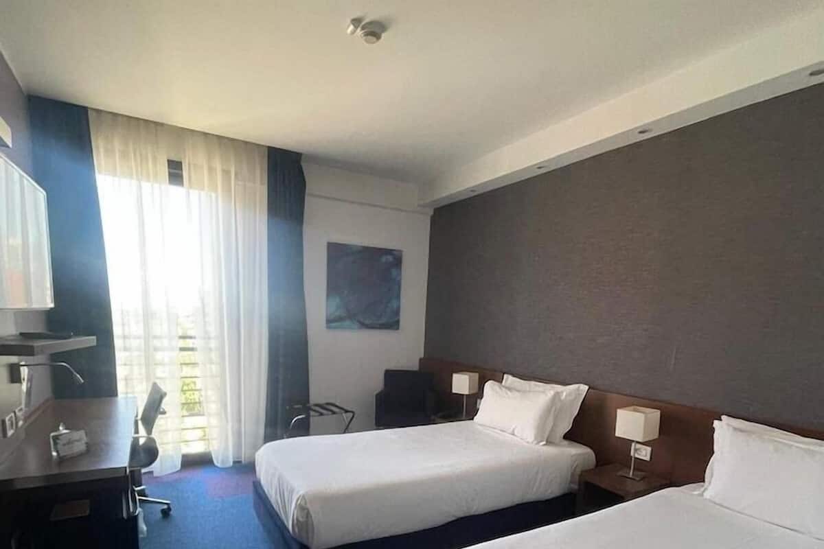 A hotel room with two beds, a desk, a chair, a TV, and a window with curtains.