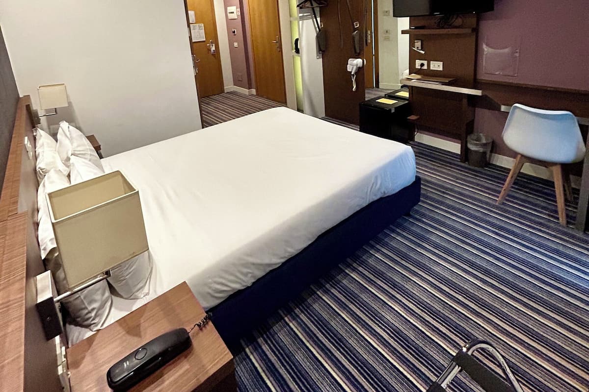 A hotel room with a large bed, a desk with a remote control, a TV, and a chair.