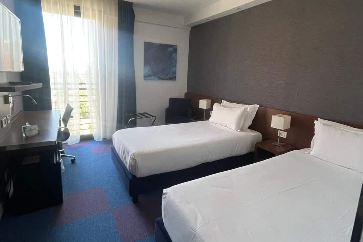 A hotel room with two beds, a desk, a chair, a TV, and a window with curtains.