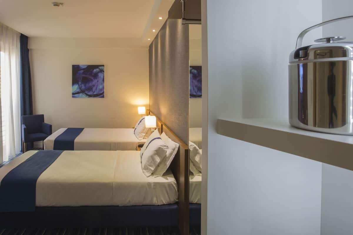 A hotel room with two beds, a desk, a chair, a painting, and a minibar.