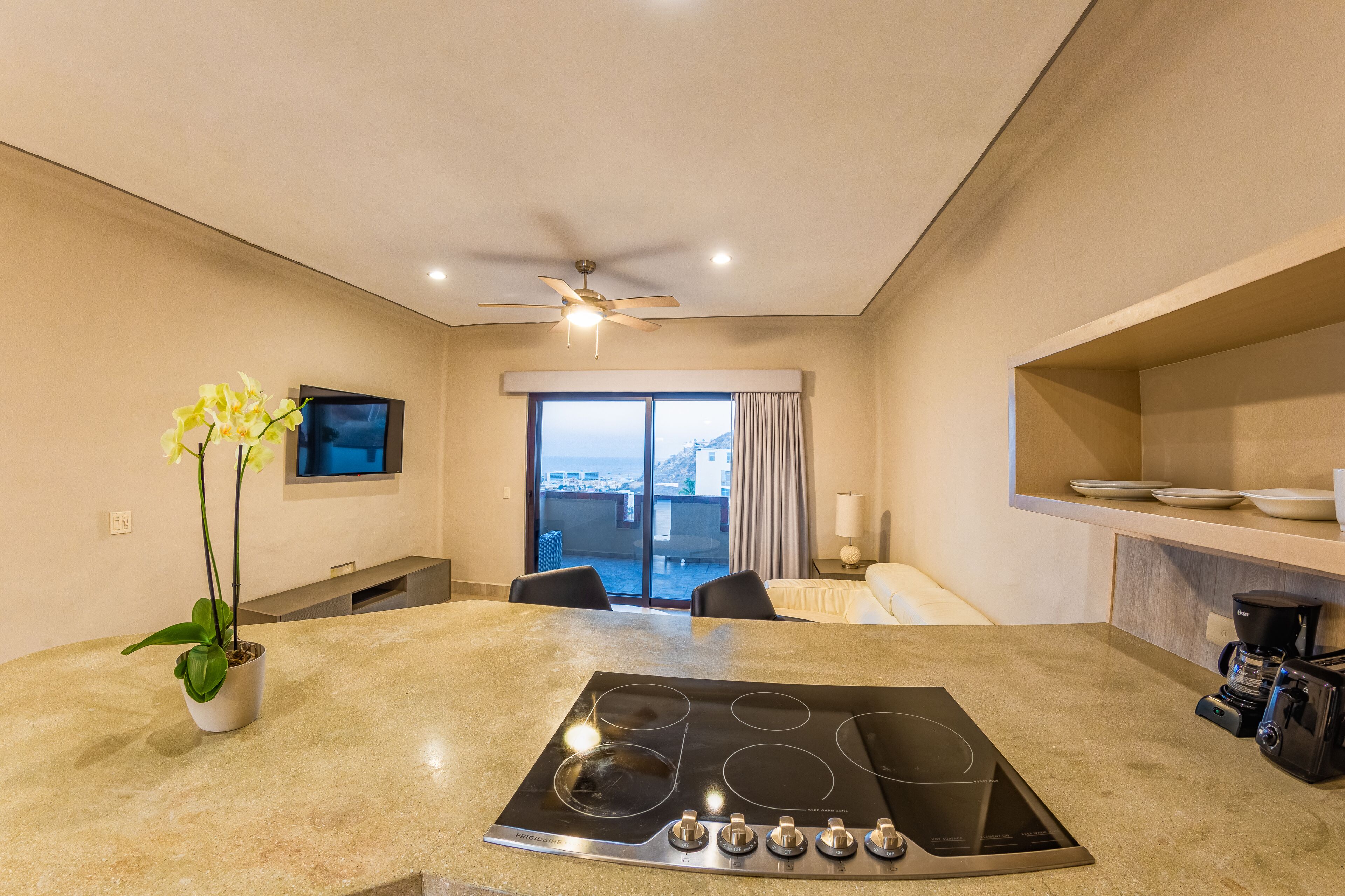 A modern kitchen with a built-in stove, a dining area with chairs, a TV mounted on the wall, and a large window with a view.