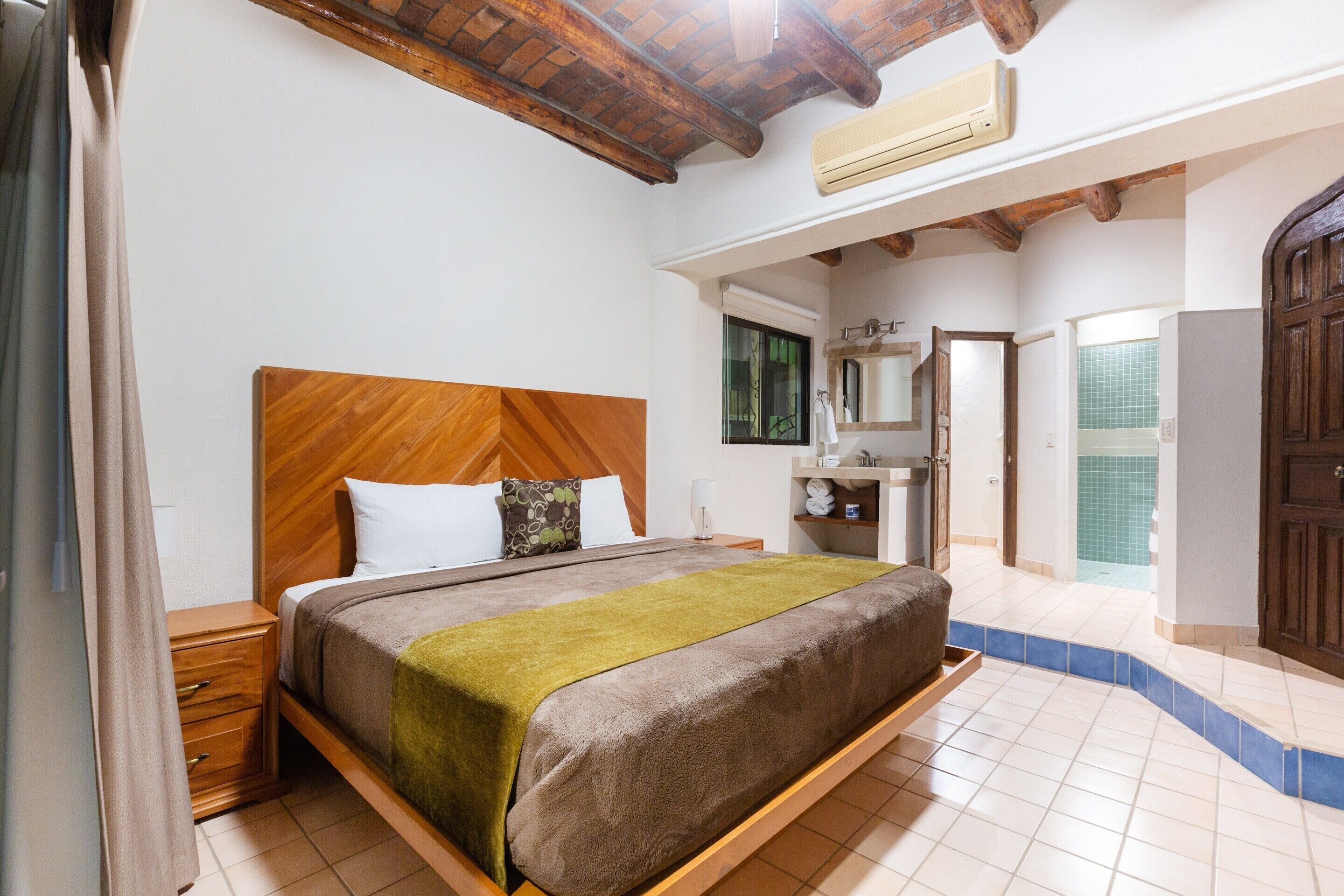 A bedroom with a wooden headboard, a bed with a green blanket, a nightstand, a dresser, a mirror, and a bathroom with a tiled floor and a wooden door.