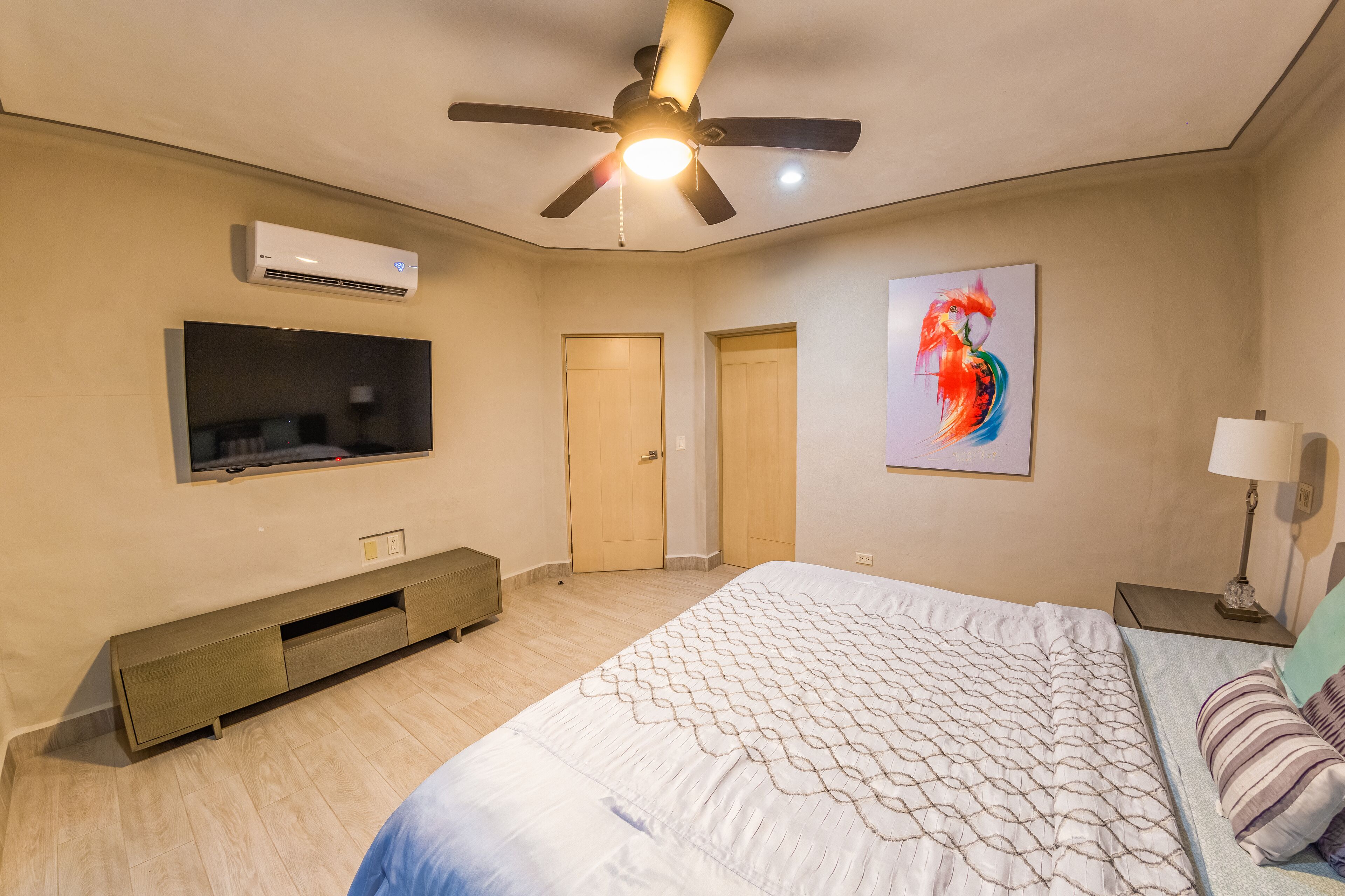 A bedroom with a bed, a ceiling fan, an air conditioner, a TV, and a painting on the wall.