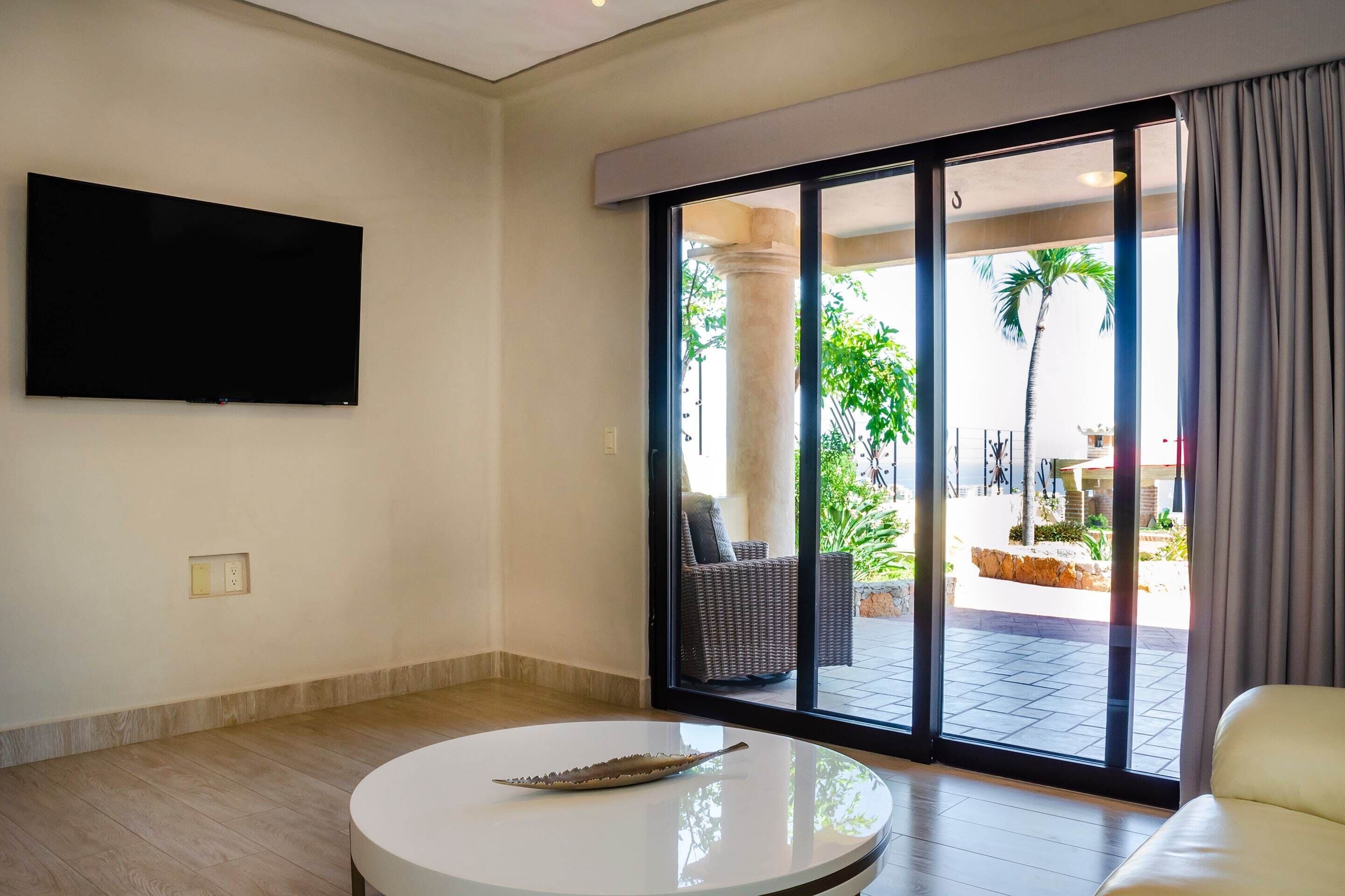 Un salon moderne avec un téléviseur à écran plat fixé au mur, une table basse ronde agrémentée d’un objet décoratif et une grande porte-fenêtre coulissante donnant sur un espace extérieur avec un palmier et des sièges.
