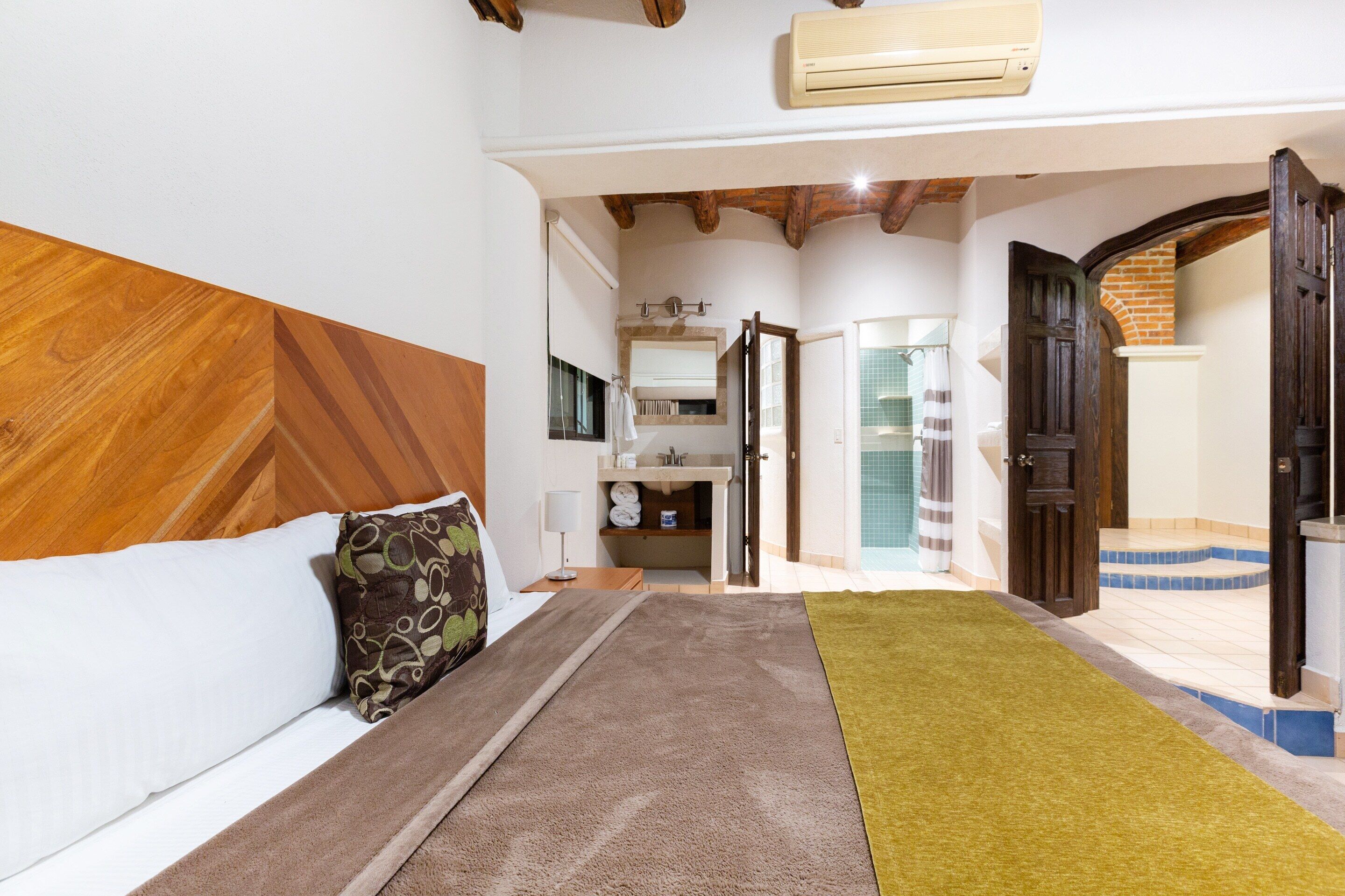 Una camera d'albergo con una testiera in legno, un letto con coperta a righe marroni e verdi, un tappeto verde e un bagno visibile attraverso una porta aperta.