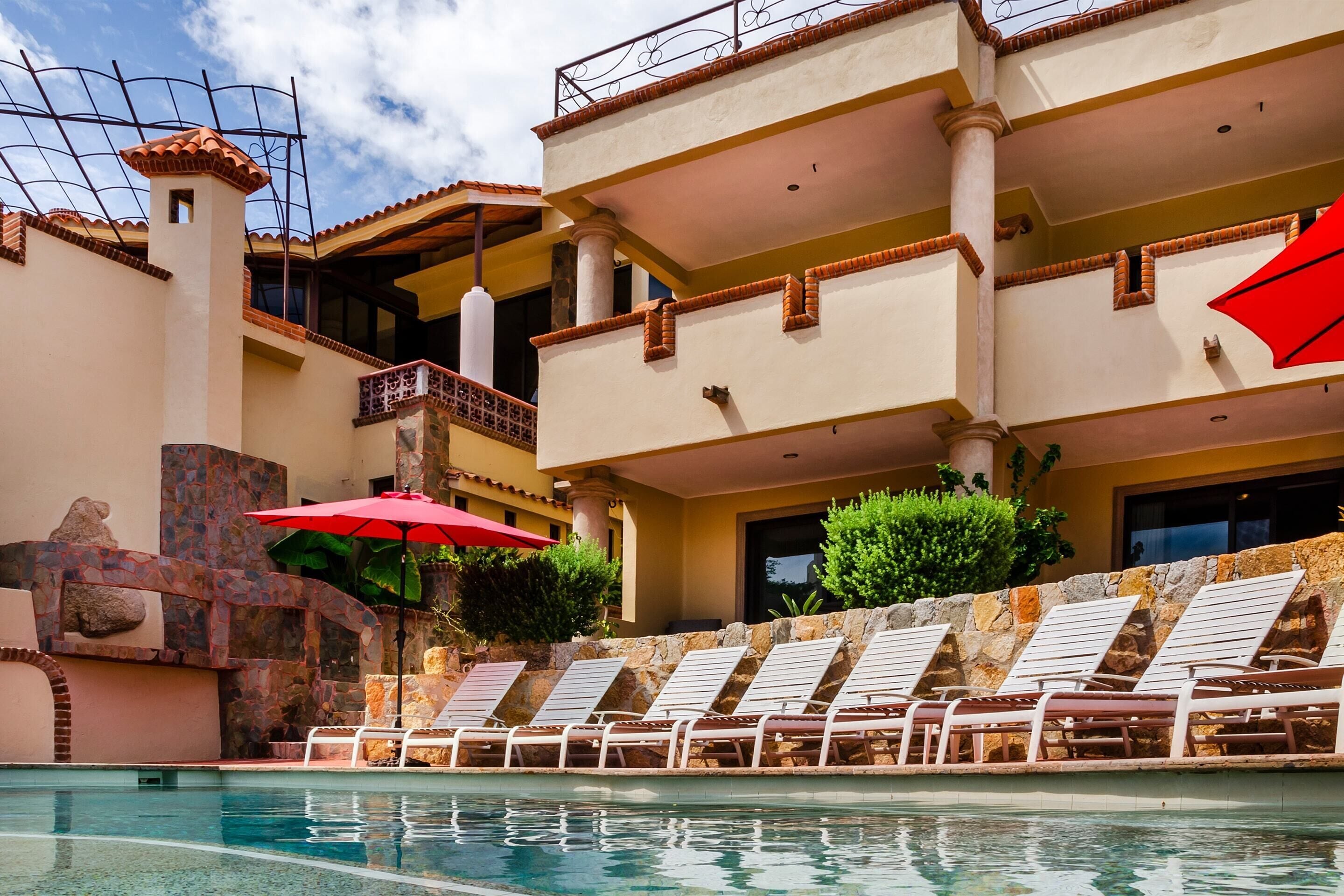 Zona de piscina con sillones de descanso y sombrillas, una pared de piedra y un edificio con varios balcones.