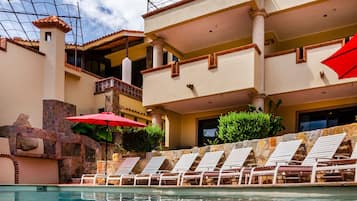 Uma área de piscina com espreguiçadeiras e guarda-sóis, um muro de pedra e um edifício com várias varandas.