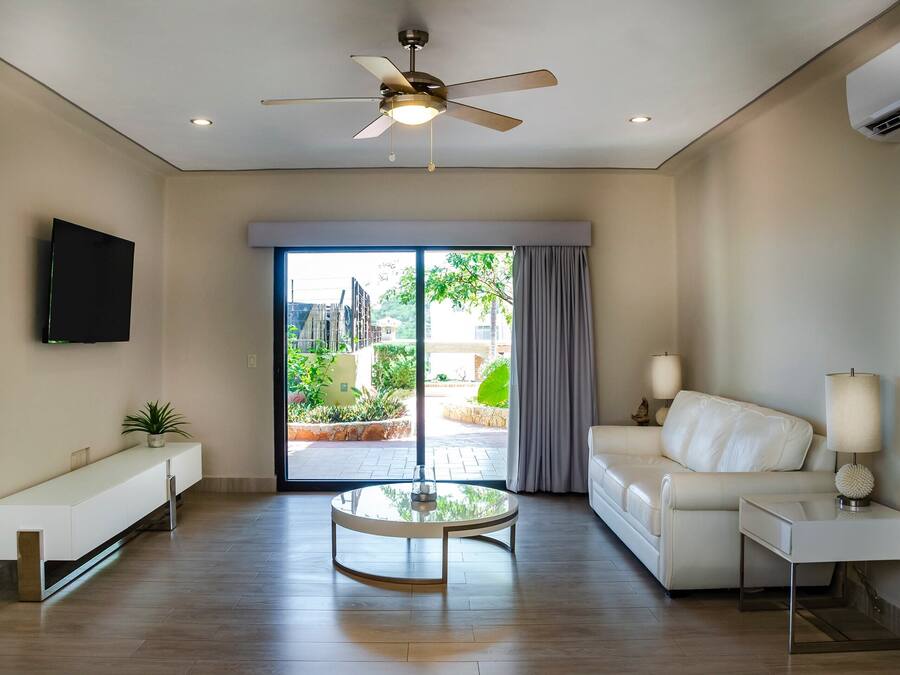 Un salon moderne avec un ventilateur de plafond, un téléviseur à écran plat fixé au mur, une table basse ronde en verre, un canapé blanc, une table d’appoint avec une lampe et un banc.