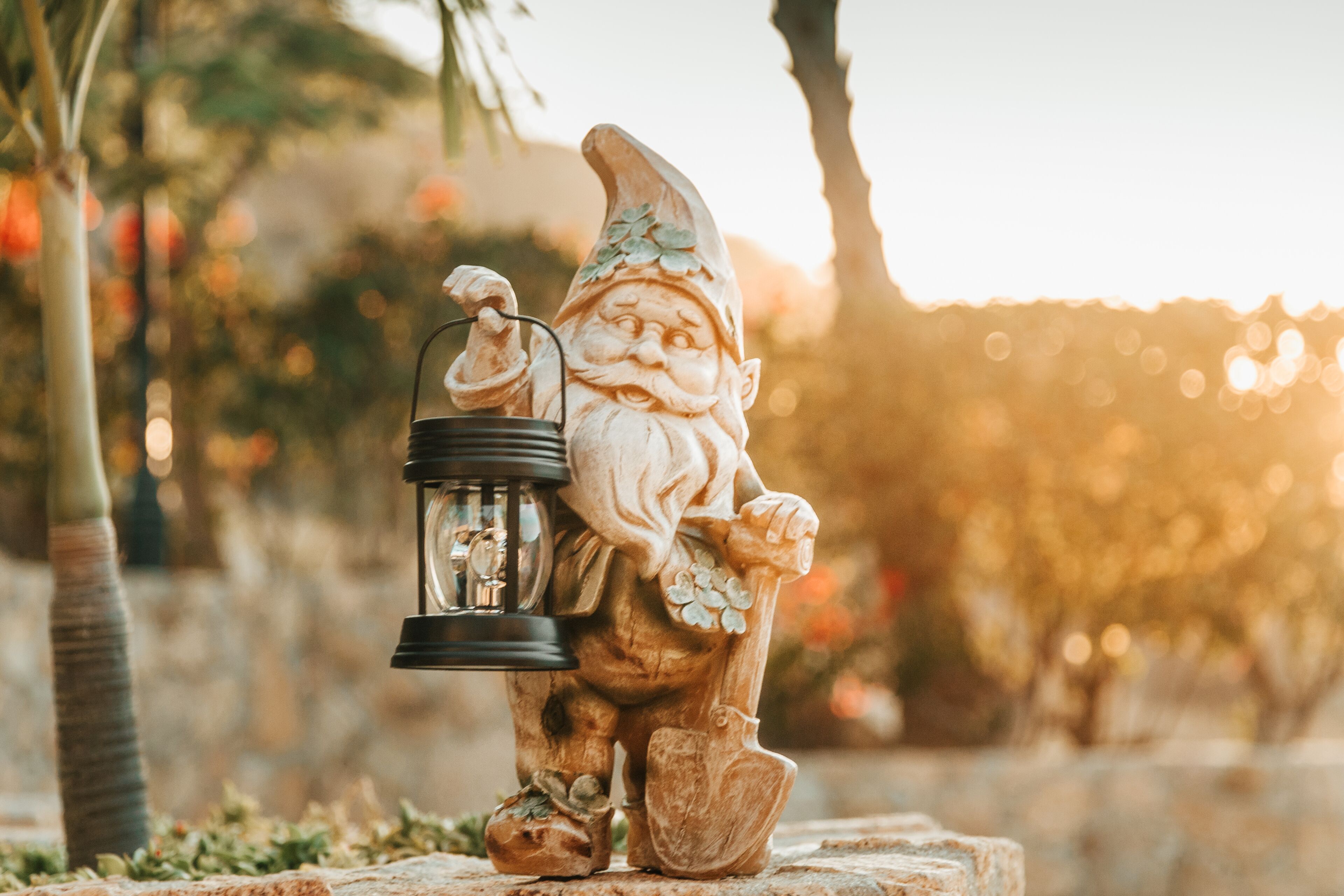 A garden gnome holding a lantern, set against a blurred background of trees and a sunset.