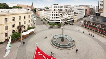 Uma praça pública com uma fonte central, rodeada por edifícios e uma bandeira.