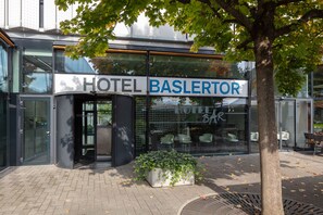 Une entrée d’hôtel moderne avec une façade en verre, une enseigne indiquant « HÔTEL BASLERTOR » et un pot de fleurs devant.