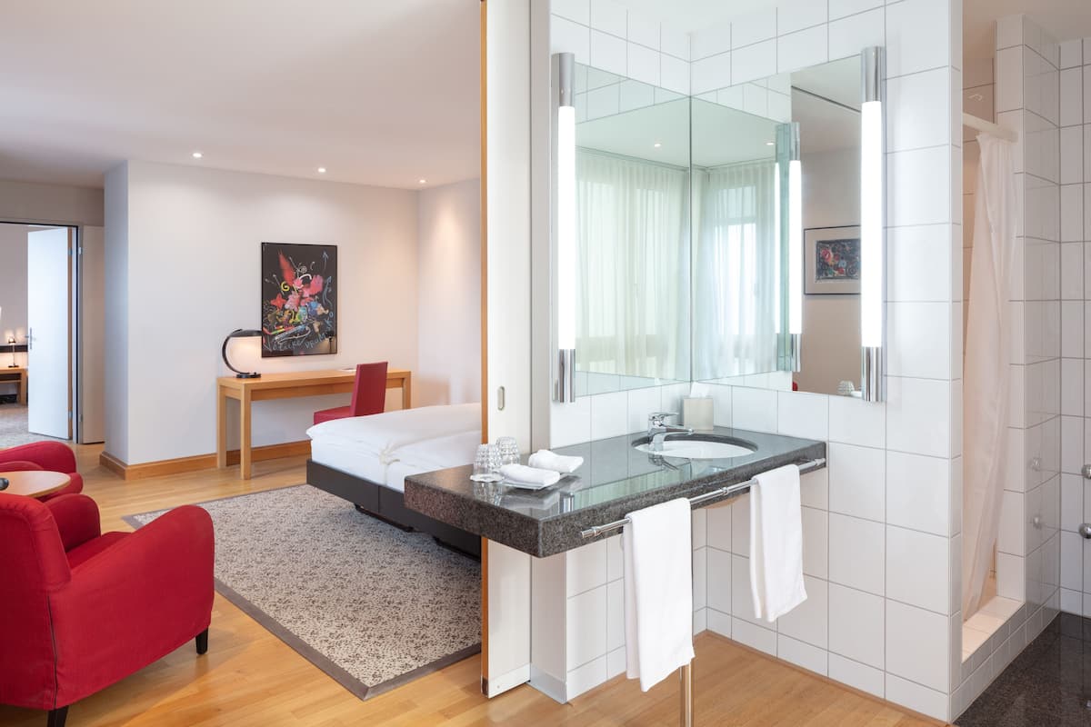 Una habitación de hotel moderna con una cama grande, una zona de estar con una mesa y sillas, un baño con ducha de vidrio y un espejo amplio.