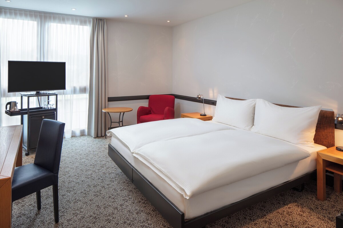 A hotel room with a bed, a desk with a chair, a TV, and a small table.