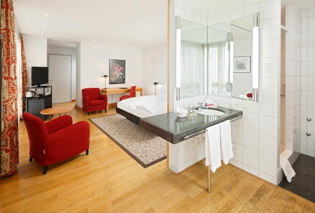 A hotel room with a bed, a sitting area with a red chair, a desk with a computer, a large mirror, and a bathroom with a sink and shower.