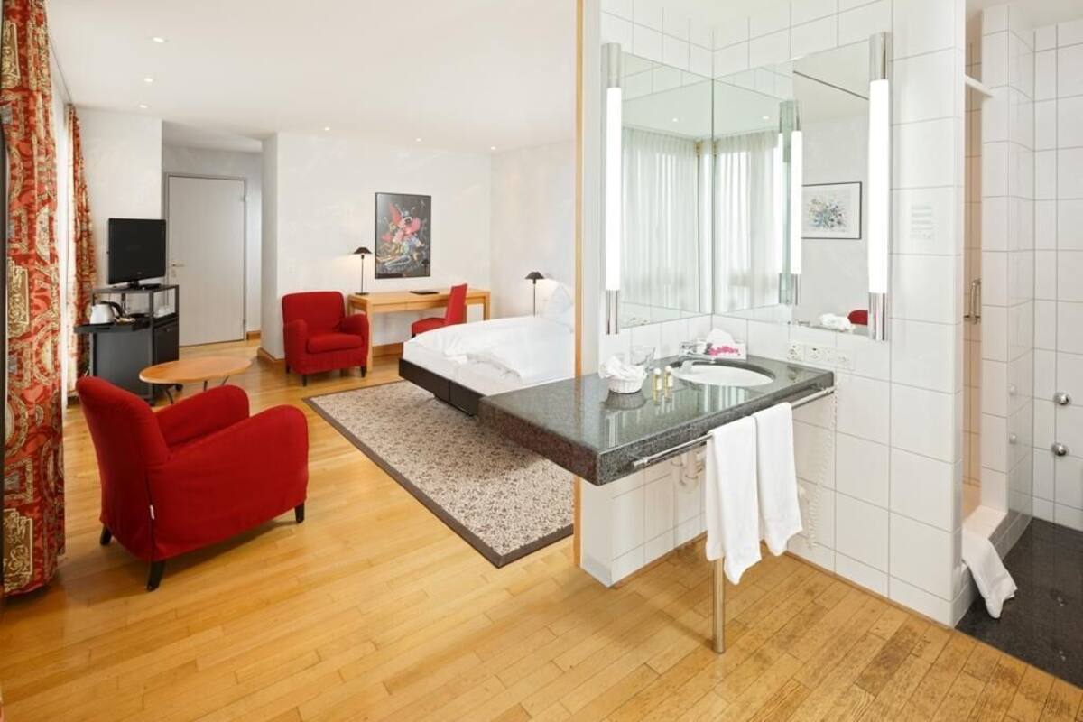 A hotel room with a bed, a sitting area with a red chair, a desk with a computer, a large mirror, and a bathroom with a sink and shower.
