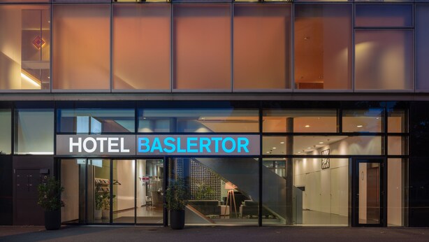 A modern hotel entrance with a glass facade, a visible staircase, and a sign reading "HOTEL BASLERTOR".