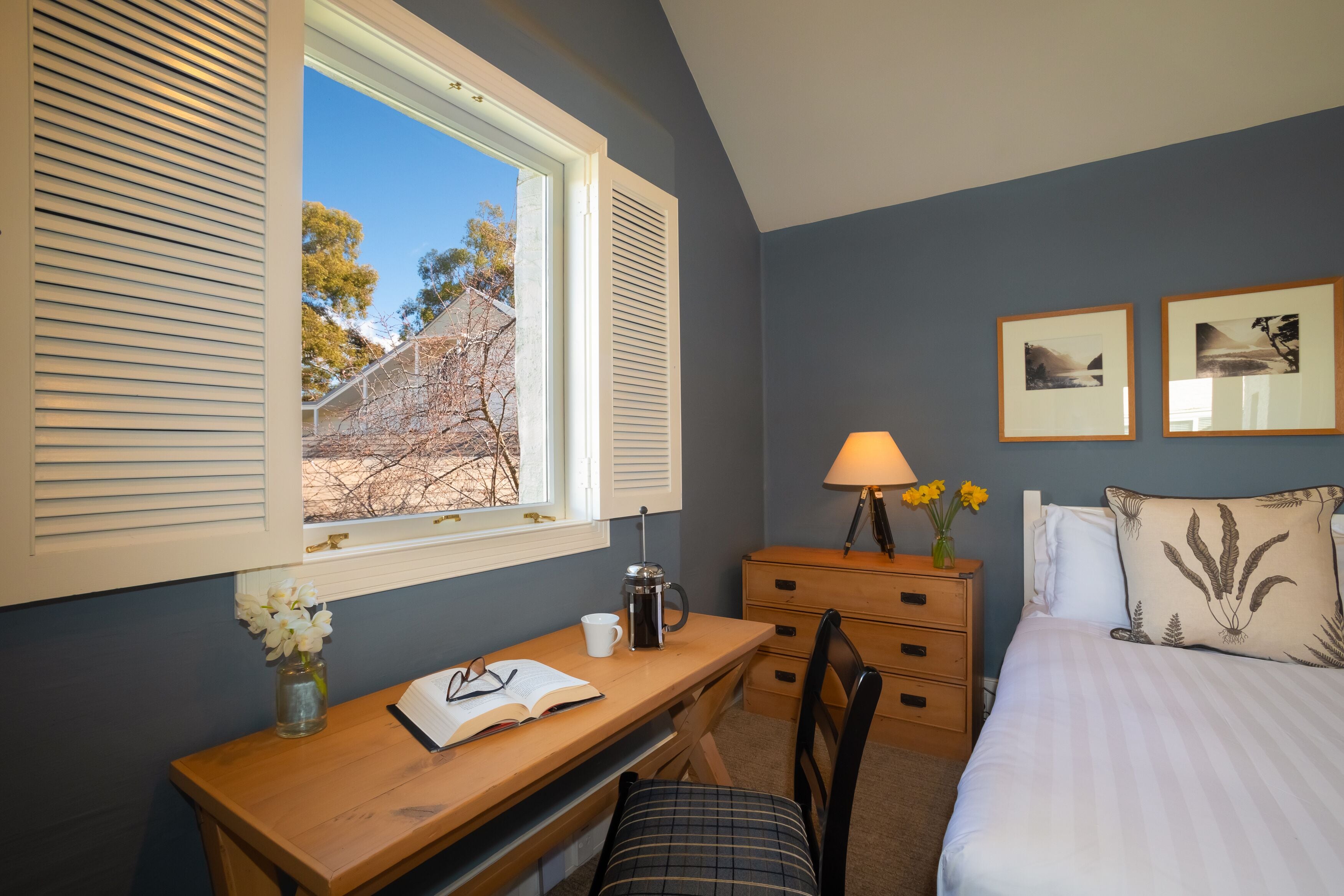 A bedroom with a bed, a desk with a lamp, a chair, a dresser, and a window with shutters.