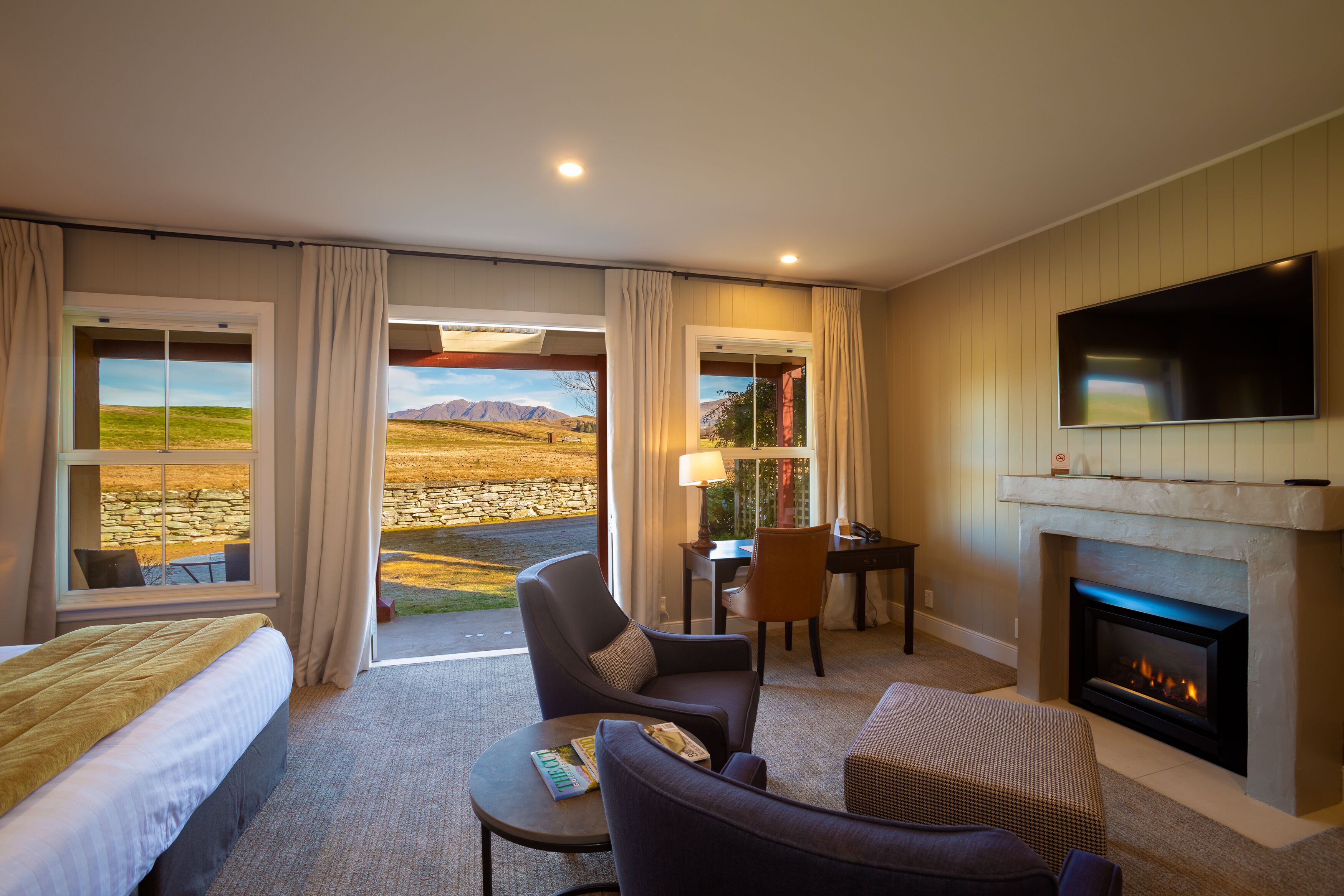 A hotel room with a bed, a seating area with a fireplace, a TV mounted on the wall, and large windows offering a view of the outdoors.