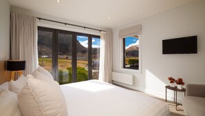 A bedroom with a large bed, a TV, a view of mountains, and a window with curtains. - Millbrook Resort (Arrowtown)