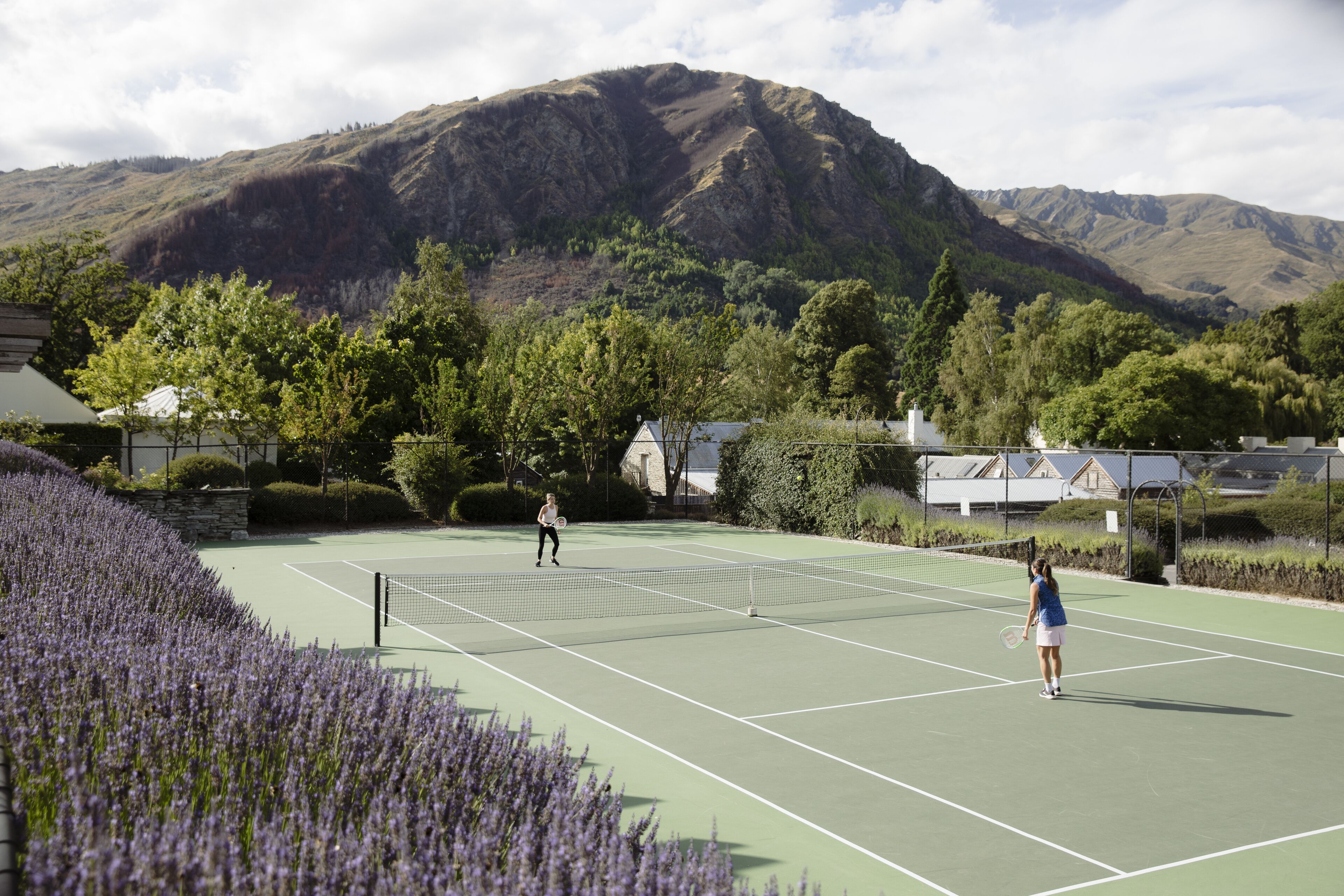 Ein Tennisplatz mit Bergelandschaft im Hintergrund, umgeben von Bäumen und einer Steinmauer.